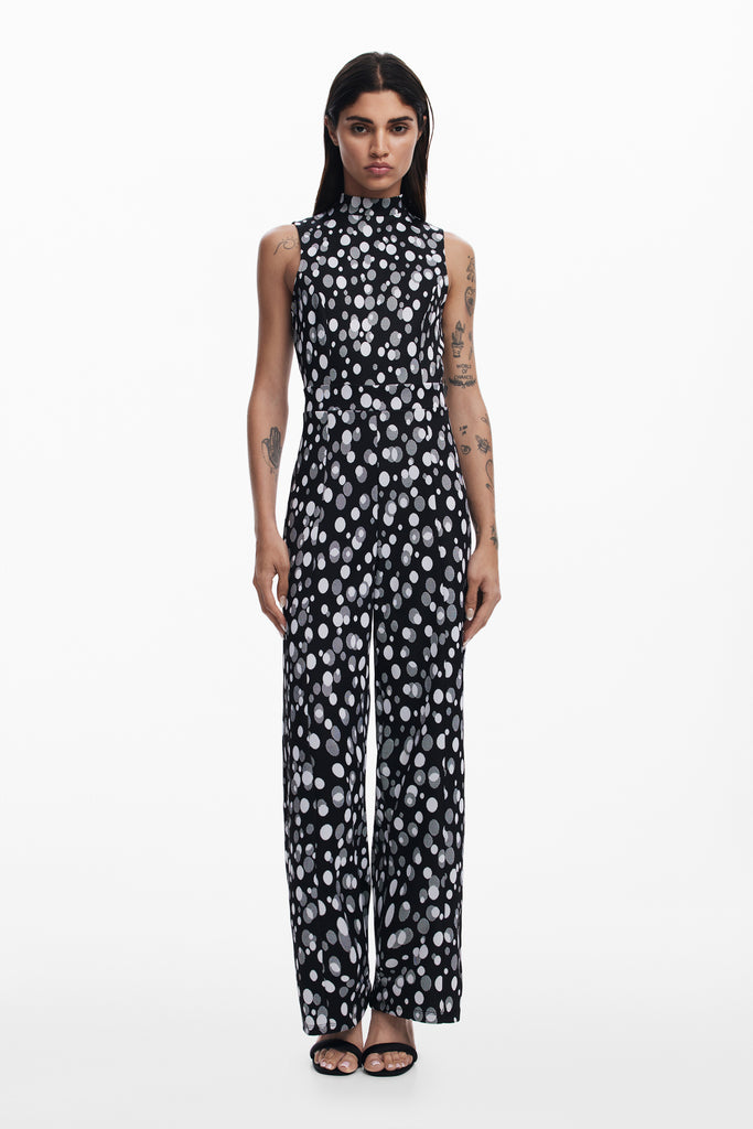 A female model wearing a sleeveless black jumpsuit with a white floral pattern, featuring a mock neck and long pants.