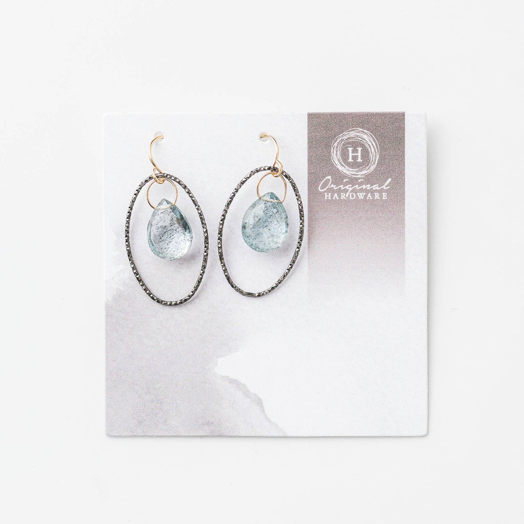 A pair of black rhodium plated sterling silver oval earrings with moss aquamarine stones, displayed against a white background with a grey logo card.