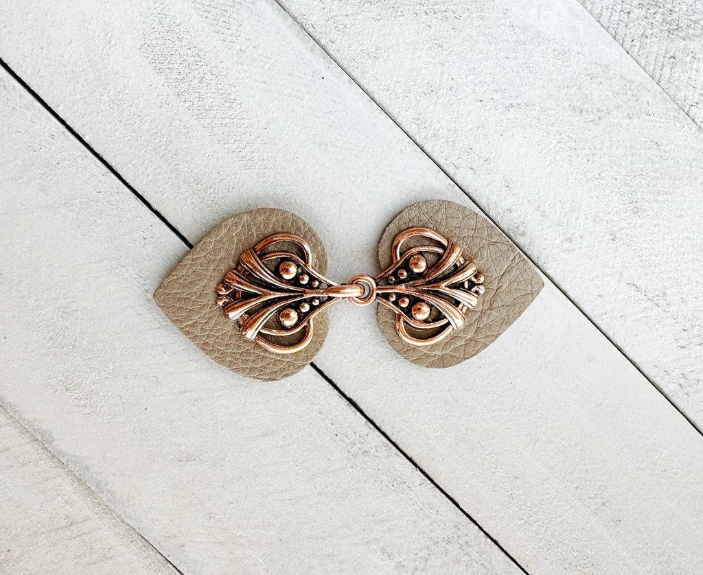 A copper cinch clip with bright hardware attached to a champagne leather strap, displayed on a white wooden background.