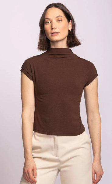 Woman wearing a brown turtleneck top and beige pants against a light purple background
