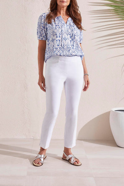 A woman standing in a white room wearing white slit ankle pants and a blue floral top, with silver sandals on her feet.