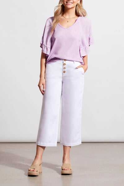 A woman is standing, wearing a purple top and white wide-leg pants with buttons at the front. She has blonde hair and is posing with her hands in her pockets.